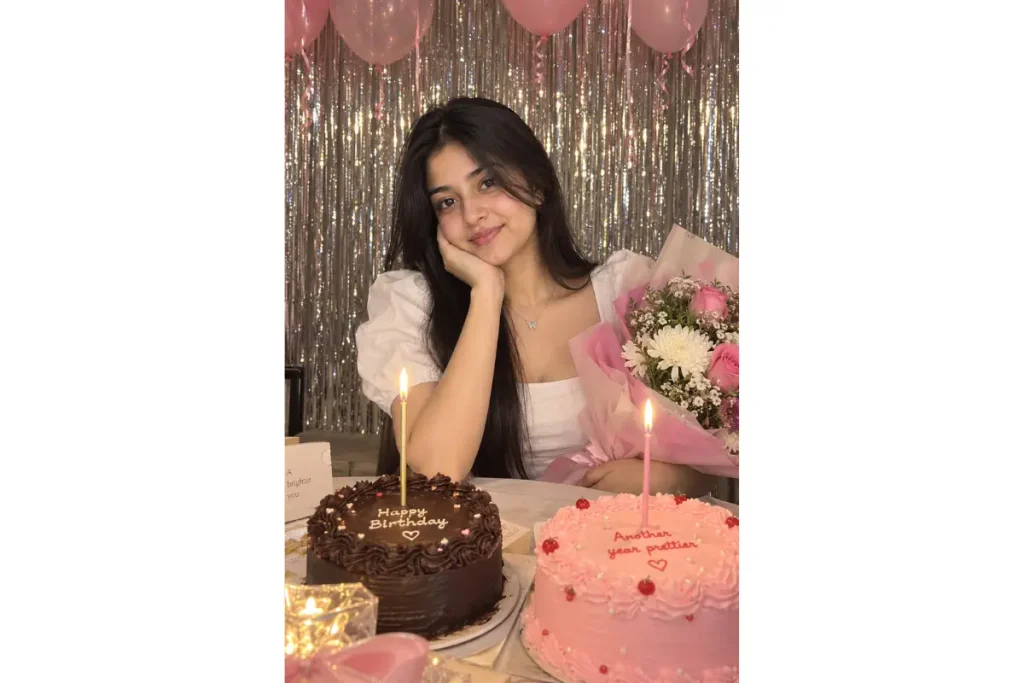 Woman posing with a pink flower bouquet and birthday cakes, with candles and a shimmering gold backdrop decorated with pink balloons.
