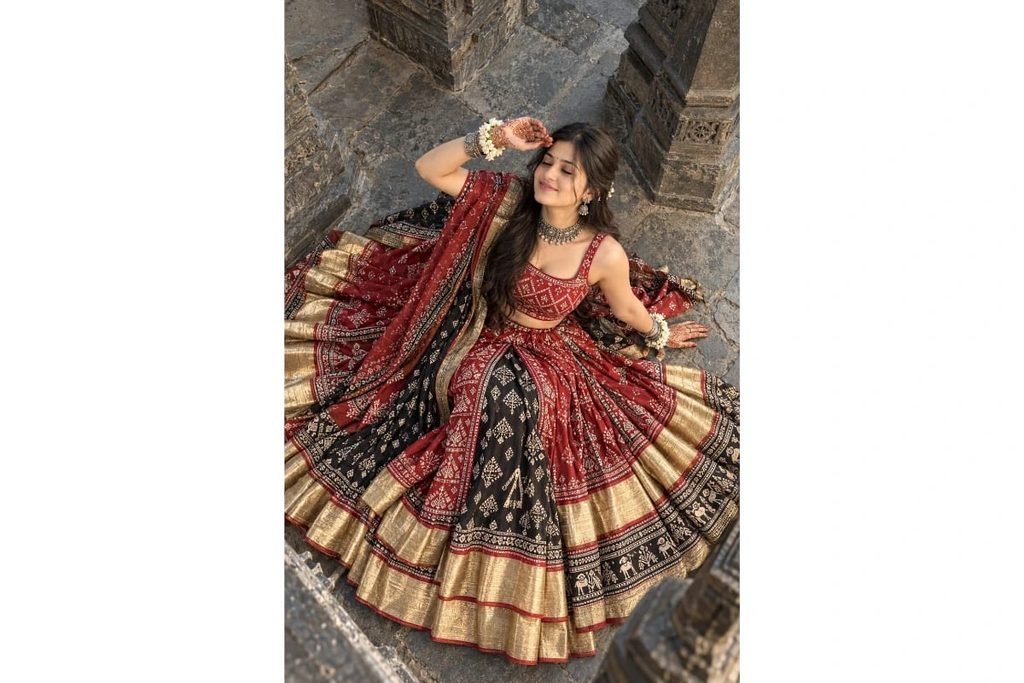 SEO-Friendly Alt Text: Red and maroon Gujarati tribal Navratri lehenga choli with traditional geometric patterns, black embroidered panels and golden borders, overhead photoshoot at an ancient stone temple