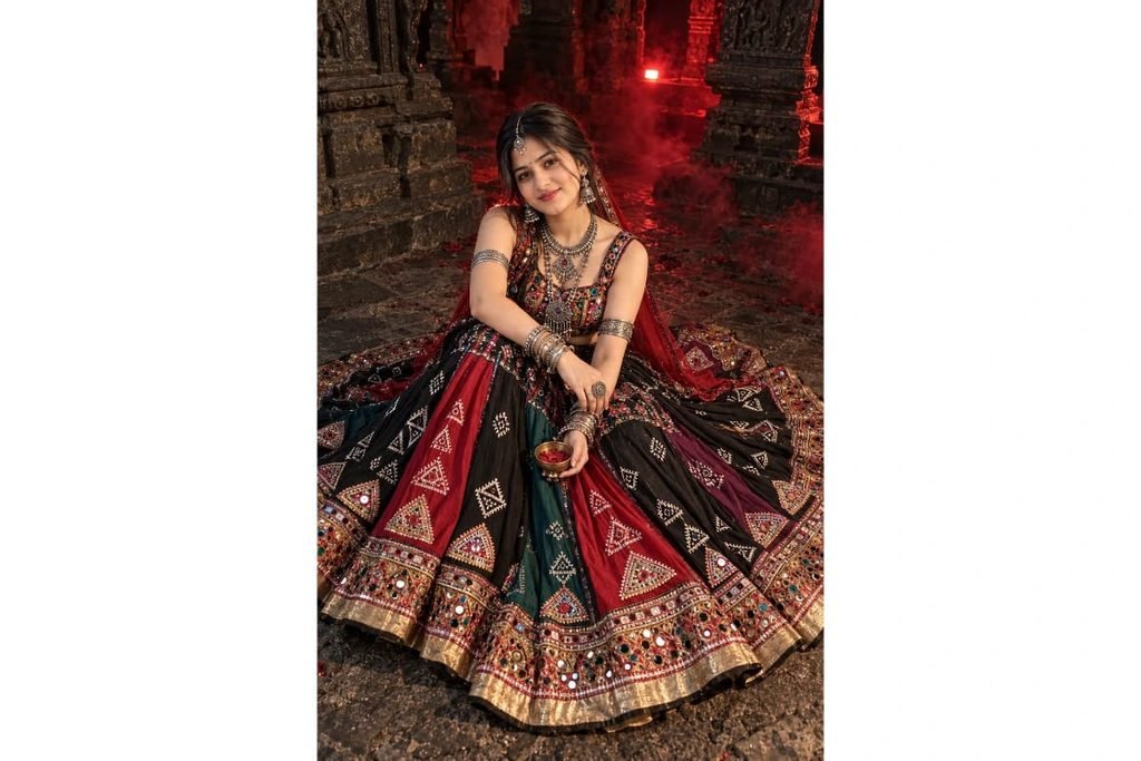 SEO-Friendly Alt Text: Dark tribal Gujarati Navratri lehenga choli with red, black and green panels, traditional geometric embroidery and golden borders, dramatic red temple lighting photoshoot for girls